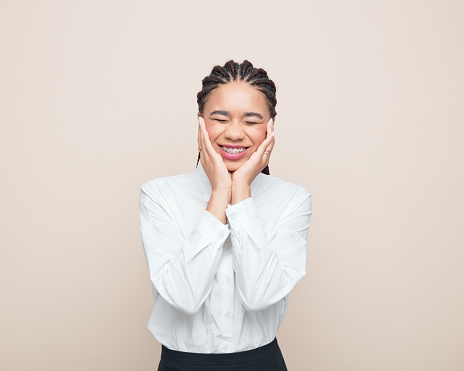 Multiracial teenage girl with braided hair wearing white shirt holding face in hands and smiling. Studio shot, beige background.