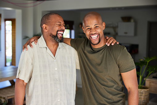 Portrait of a man and his mature father laughing while standing arm in arm together in a living room sofa at home