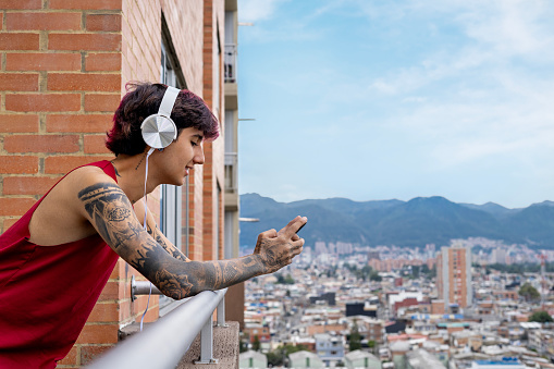 Young latin guy listening to music with headphones with his cell phone enjoying a sunny day on the balcony, in the background you can see the city, the young man is tattooed. concept music, audiobook, podcast.