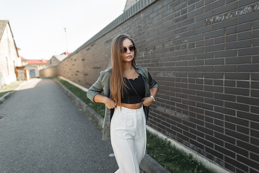 Elegant beautiful chic fashion woman with sunglasses in fashionable clothes with a shirt adjusts her pants and walks near a black brick wall on the street