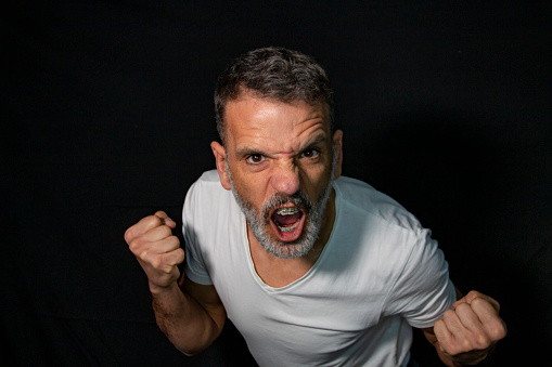 Portrait of a mature man with a gray beard, wearing braces, gesturing with his hands, wearing white t-shirt on a black background.