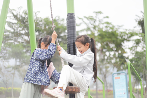 Two Asian girls are playing on the single cable pulley in the park. They help each other and cooperate, helping each other sit on the cushion of the pulley and pushing each other forward, enjoying the exciting feeling of speed.