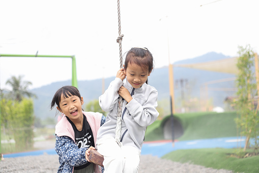 Two Asian girls are playing on the single cable pulley in the park. They help each other and cooperate, helping each other sit on the cushion of the pulley and pushing each other forward, enjoying the exciting feeling of speed.