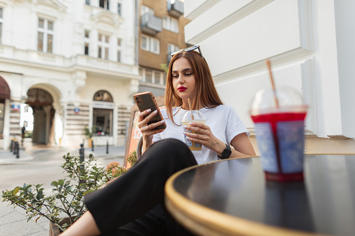 Beautiful young woman in fashion clothes sits in a cafe on the street drinking lemonade and holding a smartphone. Girl rest and reading news online