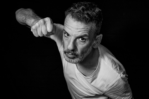 Portrait of a mature man with a gray beard, wearing braces, gesturing with his hands, wearing white t-shirt on a black background.