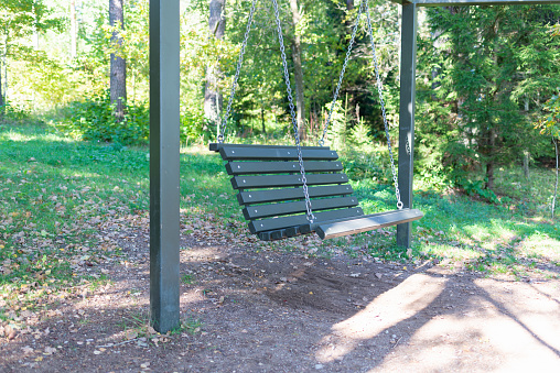 Empty hanging swing, wooden tables with benches among the trees on an autumn park, sunny day.