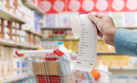 Customer showing a long grocery receipt and supermarket interior in the background, grocery shopping and budget concept