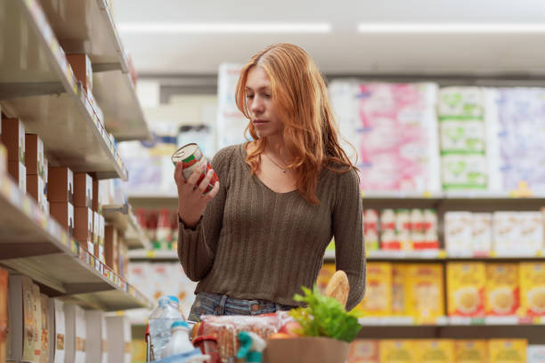 Woman doing grocery shopping and checking information on a product label Young woman doing grocery shopping at the supermarket, she is holding a can and checking information on the product label canned food stock pictures, royalty-free photos & images
