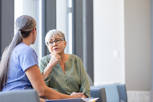 The anxious senior adult woman asks the unrecognizable female surgeon some questions as they sit in the clinic lobby.