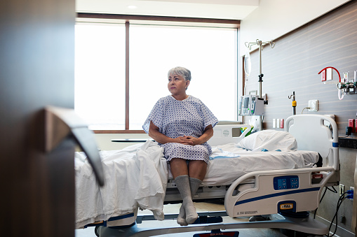 The mature female patient sits on her hospital bed and waits for the therapist to come help her walk.