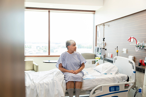 A vulnerable senior adult woman sits on the side of the hospital bed as she waits to see a doctor.
