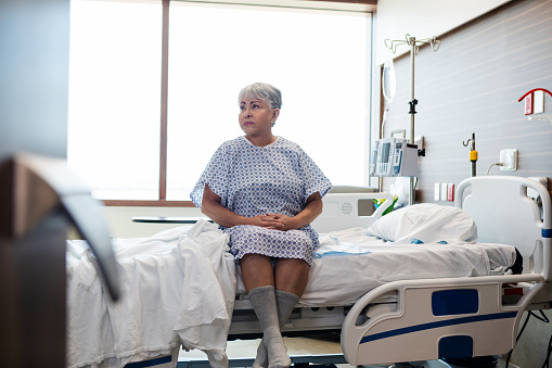 The mature female patient sits quietly on the edge of her hospital bed while she waits for the doctor.