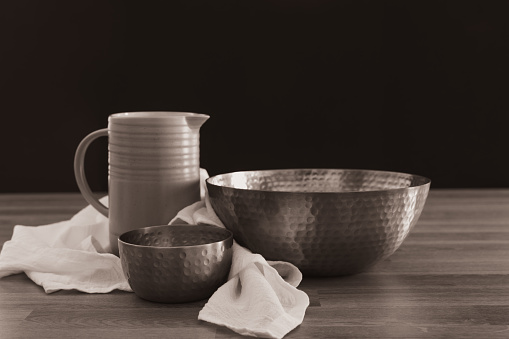 Two copper bowls of different sizes filled with water with a white linen cloth and a clay water pitcher on a light wood table shot close up with copy space and a black background