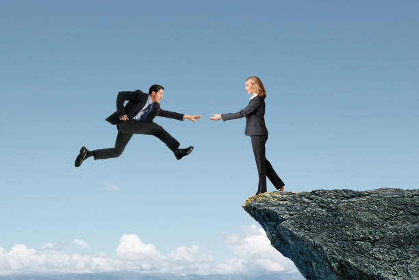 A woman standing at the edge of a cliff reaches out toward a man as he attempts to safely land.