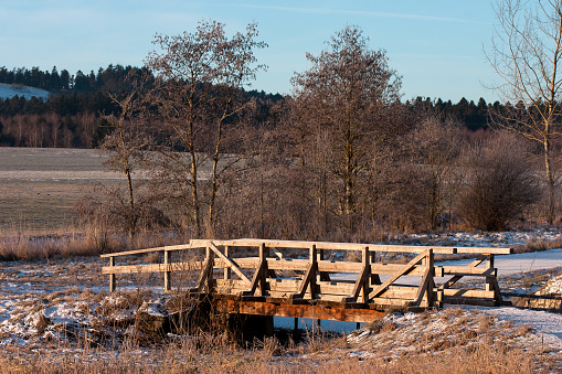 A wooden bridge spans the Schönbach between Schura and Durchhausen in winter. Lightly dusted snow, yellowed grass, create a subtle, picturesque atmosphere in the scene.