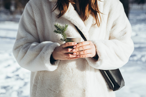 A woman in a white fur coat with a paper glass of a hot drink in the forest. Outdoors winter activities. Winter Christmas and lifestyle concept.
