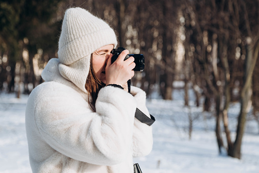 A young woman in a fur coat and hat with a camera in the winter forest, copy space. A woman photographer takes a photo in winter. Outdoor photo shoot in winter.