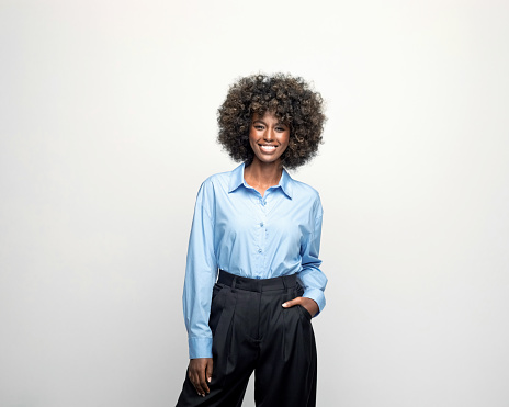 Young woman wearing blue shirt and black pants standing with hand in pocket and looking at camera. Studio shot on grey background.