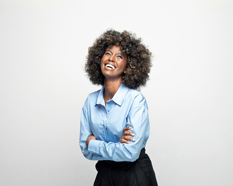 Excited young woman wearing blue shirt standing with arms crossed, looking up and laughing. Studio shot on grey background.