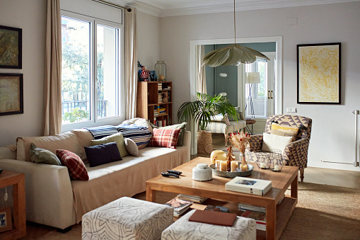 Unoccupied living room with sofa, coffee table, ottomans, hanging lamp, armchair, potted plant, knick knacks, and natural daytime window light.