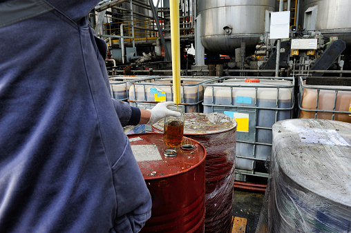 A worker is obtaining a sample from a red drum at an industrial facility.