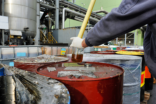 A worker wearing protective gloves draws a liquid sample from an industrial barrel.