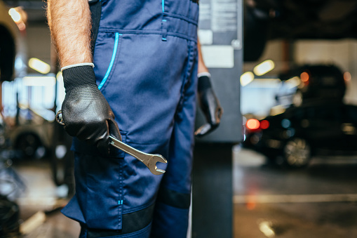 Car repairman wearing a dark blue uniform standing and holding a wrench with vehicle on blurred background.
