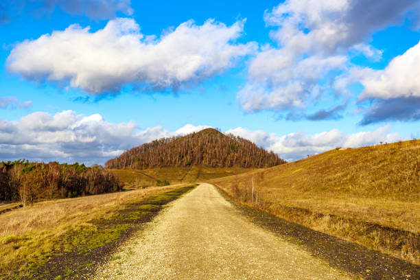 Hiking path between plain and hills with a mountain with autumn trees against the blue sky with white clouds in the background, Thor Park - Hoge Kempen National Park, sunny afternoon in Genk, Belgium
