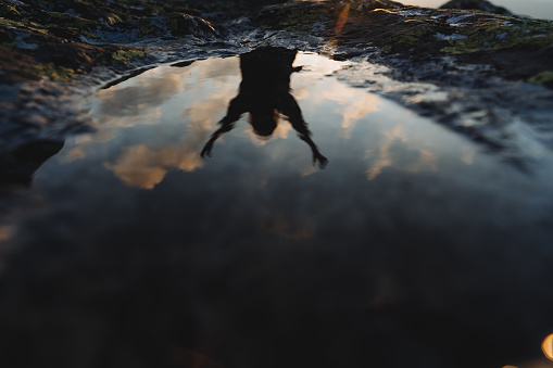 Reflection of the silhouette of a man in lying on a stone in the mountains, morning dawn, man raising his hands up, inverted image, sky reflected in water. High quality photo