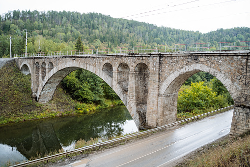 An ancient arch bridge over the river, an architectural structure of the 19th century, a railway bridge, a road runs along the river, a landscape in a mountainous area. High quality photo