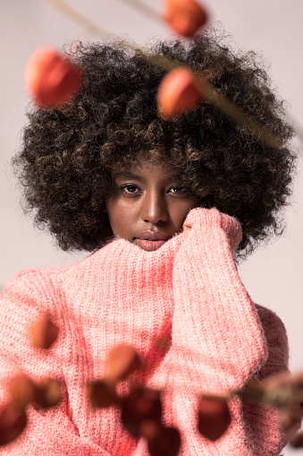 Headshot of young woman with afro hairstyle wearing pink wool sweater, looking at camera among twig of orange physalis.