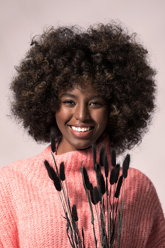 Headshot of young woman with afro hairstyle wearing pink wool sweater, holding dry grass, looking away and smiling.