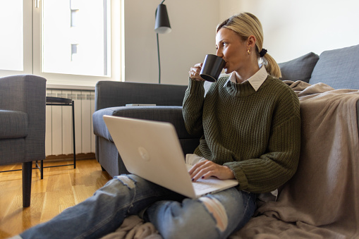 A young woman sitting on the floor of her living room, drinking coffee, maybe taking a break from work. A laptop is on her lap.