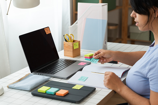 A woman sits at a desk, using her laptop and consulting colorful sticky notes for productivity.