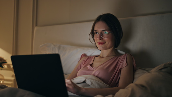 Smiling woman finishing work in night bed. Happy girl resting closing computer in evening bedroom. Satisfied late manager freelancer browsing web making project. Joyful relaxed girl going to sleep