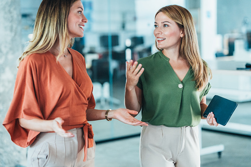 Shot of a two confident business female colleagues chatting walking through the office hall, enjoy friendly warm conversation. Business women in the office having informal talk in coffee break. Smiling business persons having a conversation walking and talking