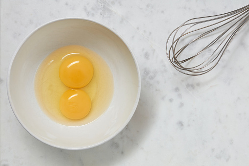 Double yolk eggs in a bowl and wire whisk on a white marble surface. Two yolks in one chicken egg. Identical twins. Double eggs. Flat lay. Copy space