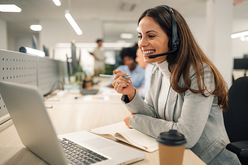 Smiling caucasian business woman with headset working on a laptop in a call center on the line. Happy female working in a busy customer service department and taking notes while talking with a client.