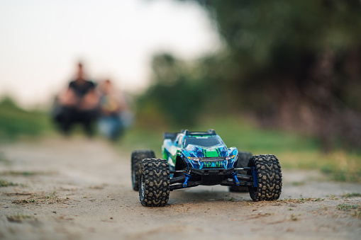 Selective focus on a toy electric car, remotely controlled on a dirt road in the countryside. Blurred background shows two figures, a boy and his father, that are operating the electric toy car.