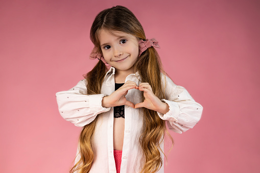 Adorable little girl making a heart with her hands, smiling at the camera, isolated on a pink background. Valentine's day concept. High quality photo