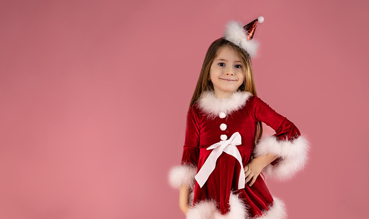 A cute little girl with long wavy blond hair in a red dress and head accessory is smiling beautifully. Pink background, space for text. High quality photo