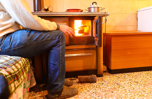 Elderly person sitting next to the old wood stove of the economical kitchen with the fire lit