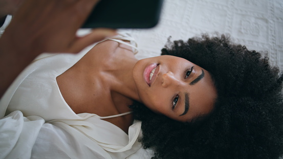 Peaceful girl read message in bed alone. Calm african american lady laying bedroom watching smartphone movie at home. Curly hair model relaxing at soft hotel mattress. Dreamy woman resting at weekend