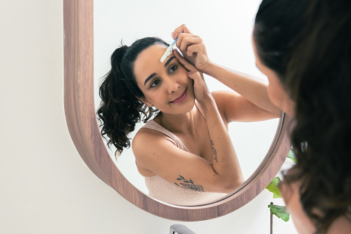 Close up shot of Latin American female looking at a mirror for observing her eyebrow and face skin before applying cosmetics on her face at a bathroom. Young adult woman having beauty treatment activity at home.