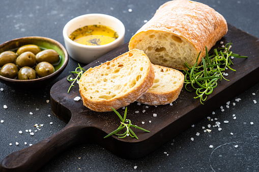 Ciabatta bread on wooden board with olive oil, olives and herbs on black. Mediterranean food.