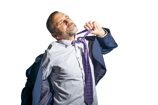 Madrid, Spain. Half-length view of a stressed mature bearded man in a suit and tie, feeling overwhelmed by his attire, attempting to relieve himself by loosening his workwear. Isolated against a white background.