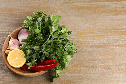 Bowl with fresh green parsley, chili peppers, lemon, onion and garlic on wooden table, above view. Space for text