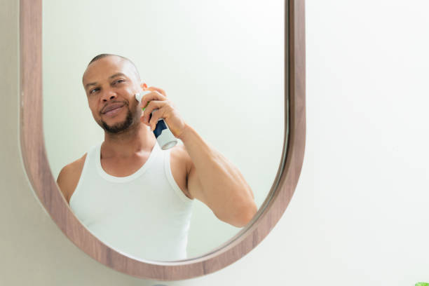 happy and handsome latin american male applying shaving cream on his face to clean his beard during standing in front of mirror in bathroom. mid adult black man shaving his facial hair in morning. reflection in the mirror image. - shaving men electric razor reflection imagens e fotografias de stock