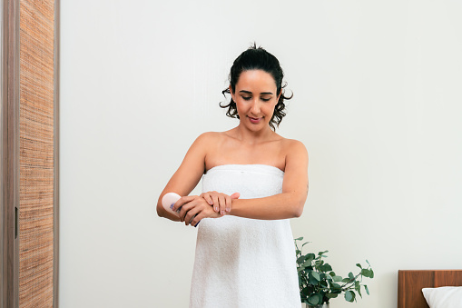 Beautiful Latin American female wearing white towel, applying moisturizing lotion for dry skin to improve soft and flawless skin on her arms after taking a bath in bedroom. Beauty routine. Daily skincare
