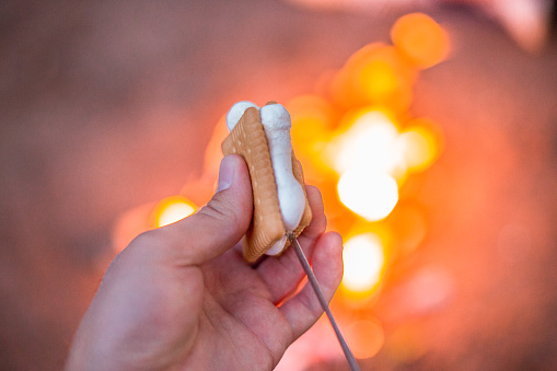 Close up of hands holding S'more made with marshmallow and cookies, in the background fire